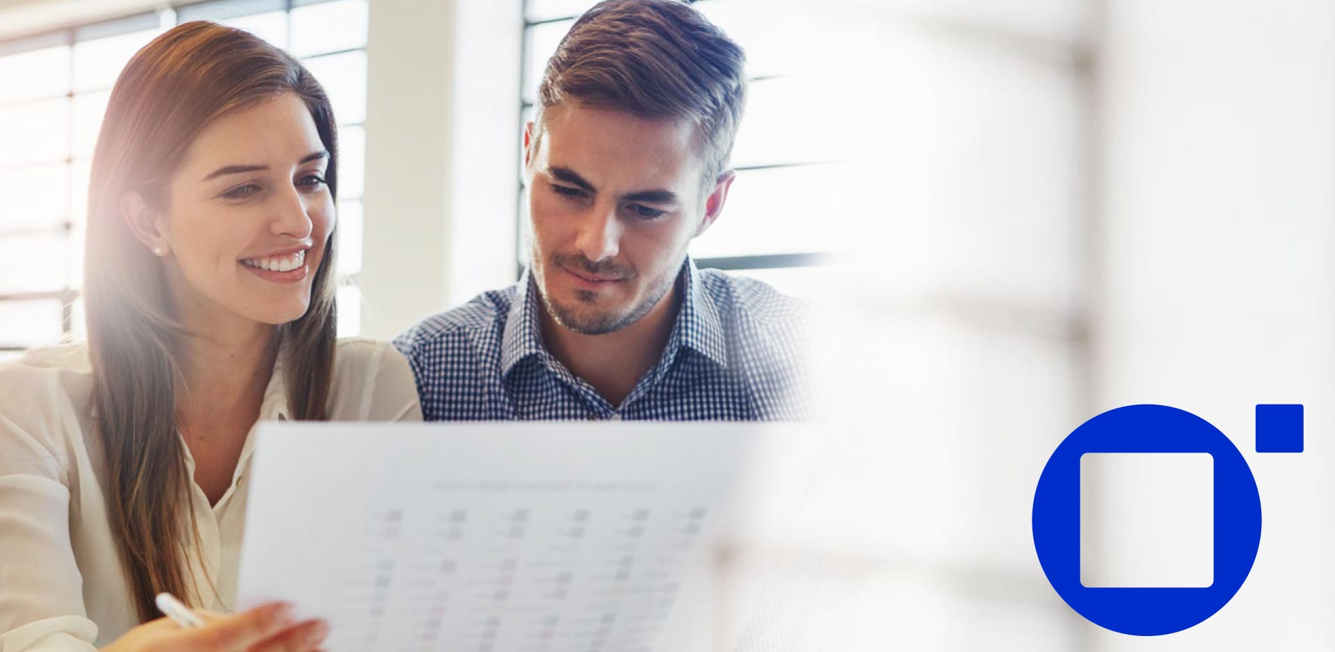 Blurred image of two people sitting side by side reviewing a document, with a large blue rounded-square Syspro-style logo on the right against a bright, softly lit office background.