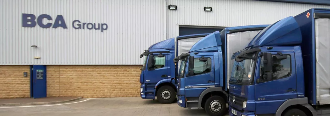 Three blue delivery trucks parked outside a BCA Group warehouse building.