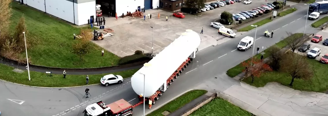 Aerial view of a large industrial tank being transported on a long multi-axle trailer outside an engineering facility, with workers, vehicles, and onlookers nearby