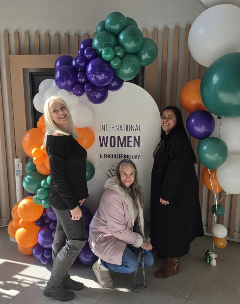 Three Syspro colleagues pose at the International Women in Engineering Day balloon display in the office lobby.